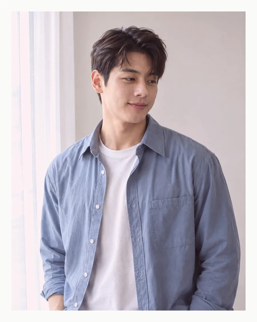 흰 여백 안 흰 커튼 곁에서 하늘색 셔츠 소매를 걷고 선 모습